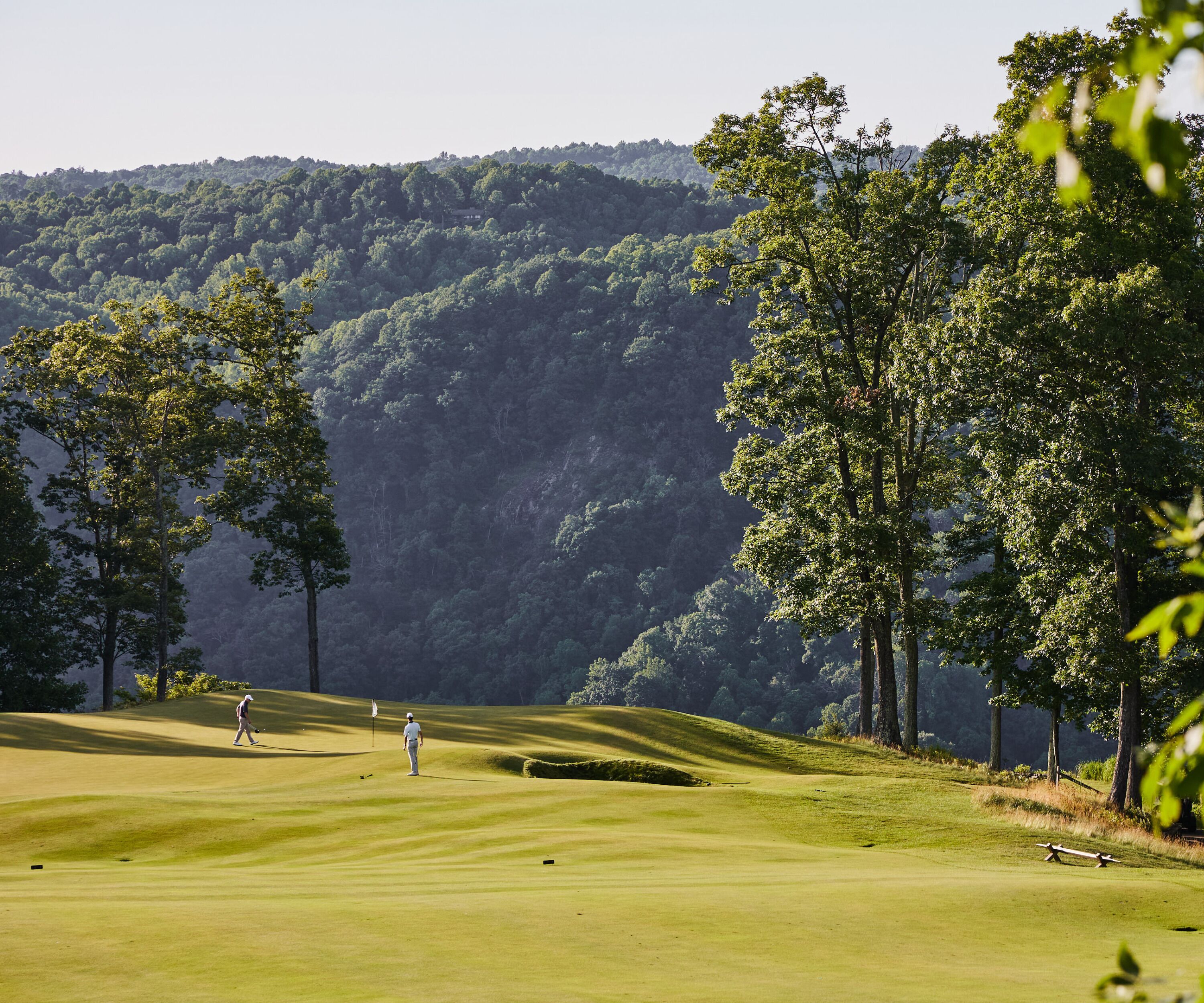 A golf course with a person walking on the fairway, surrounded by trees and hills in the background.