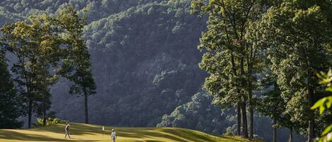 Un campo de golf con una persona caminando por el fairway, rodeado de árboles y colinas al fondo.
