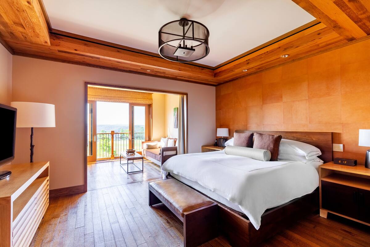 A spacious bedroom with a large bed, wooden ceiling beams, a balcony, and a television on a wooden stand.