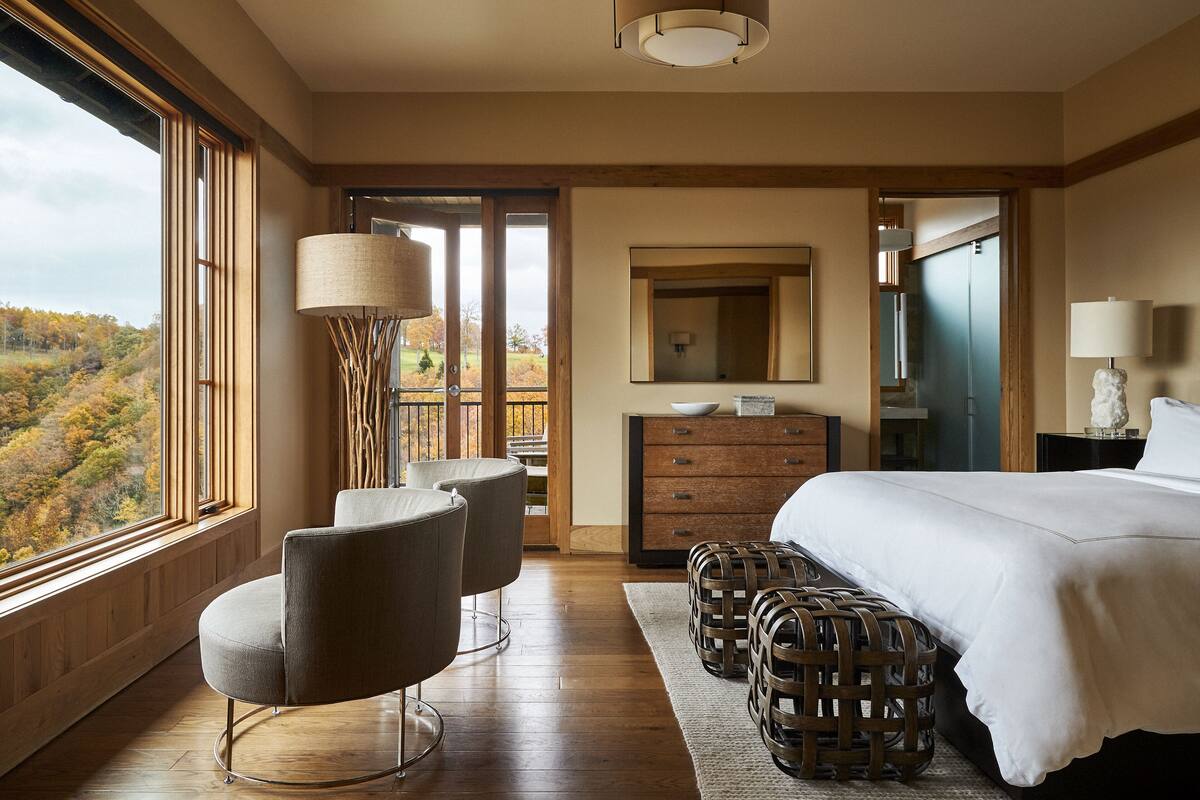 A bedroom with a large bed, a dresser, a mirror, and a view of a forest through a large window.