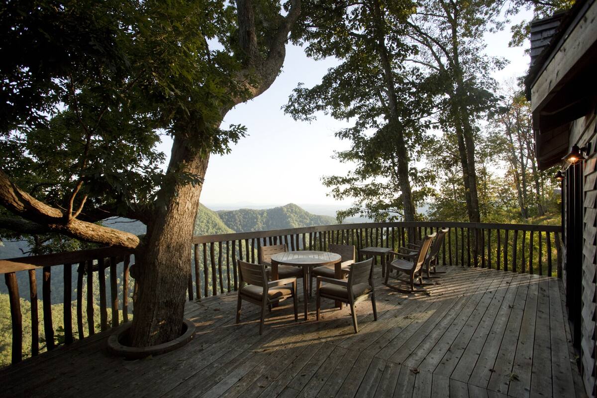 A wooden deck with a round table and chairs, surrounded by a wooden railing, overlooking a mountainous landscape.