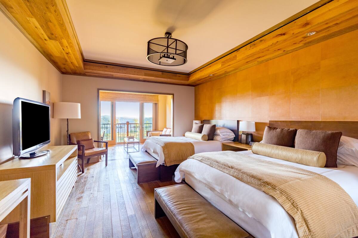 A hotel room with two beds, a desk with a TV, a chair, and a balcony view.