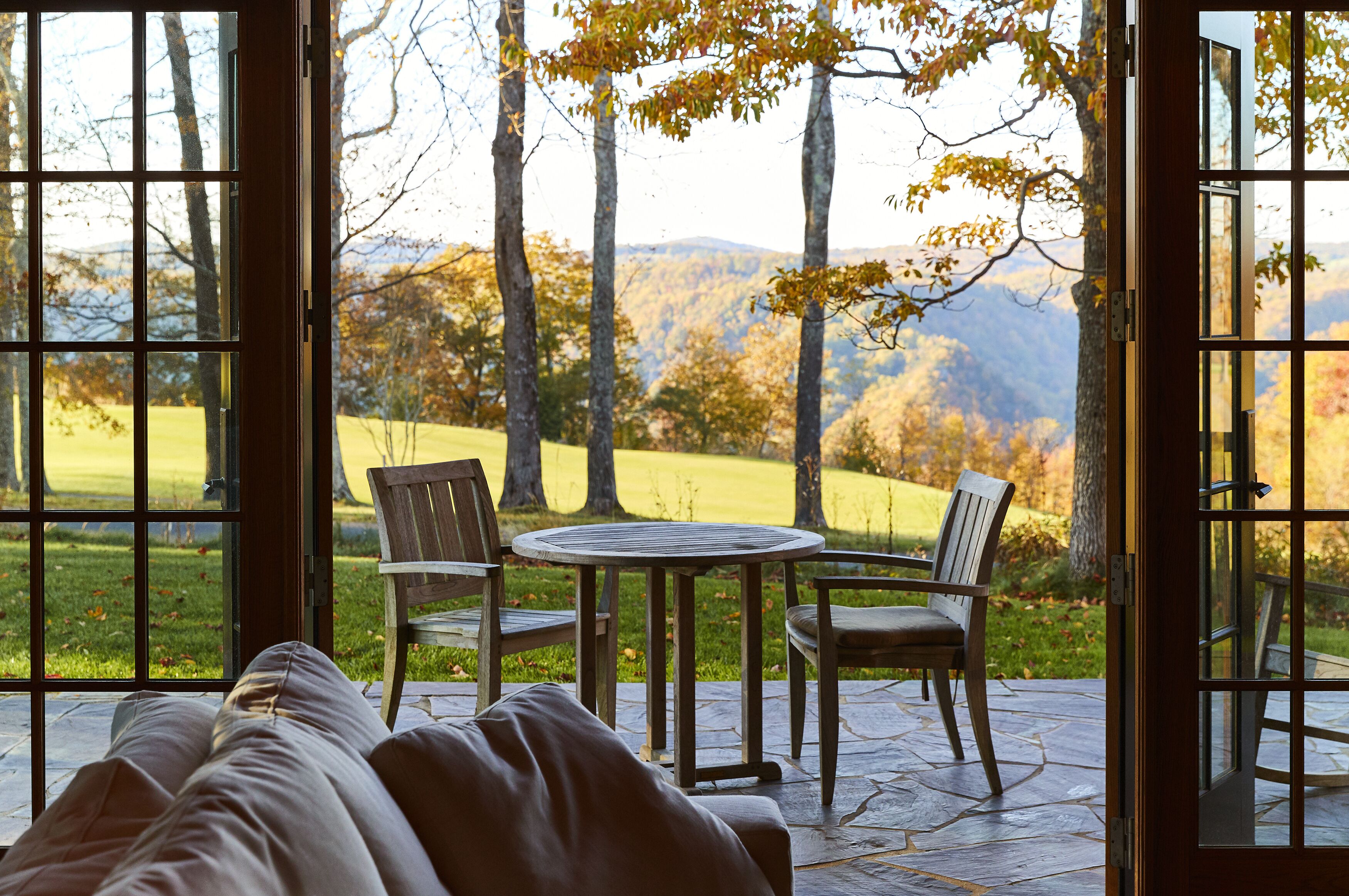 A room with a large window offering a view of a grassy field and trees. There is a round table with two chairs and a sofa.