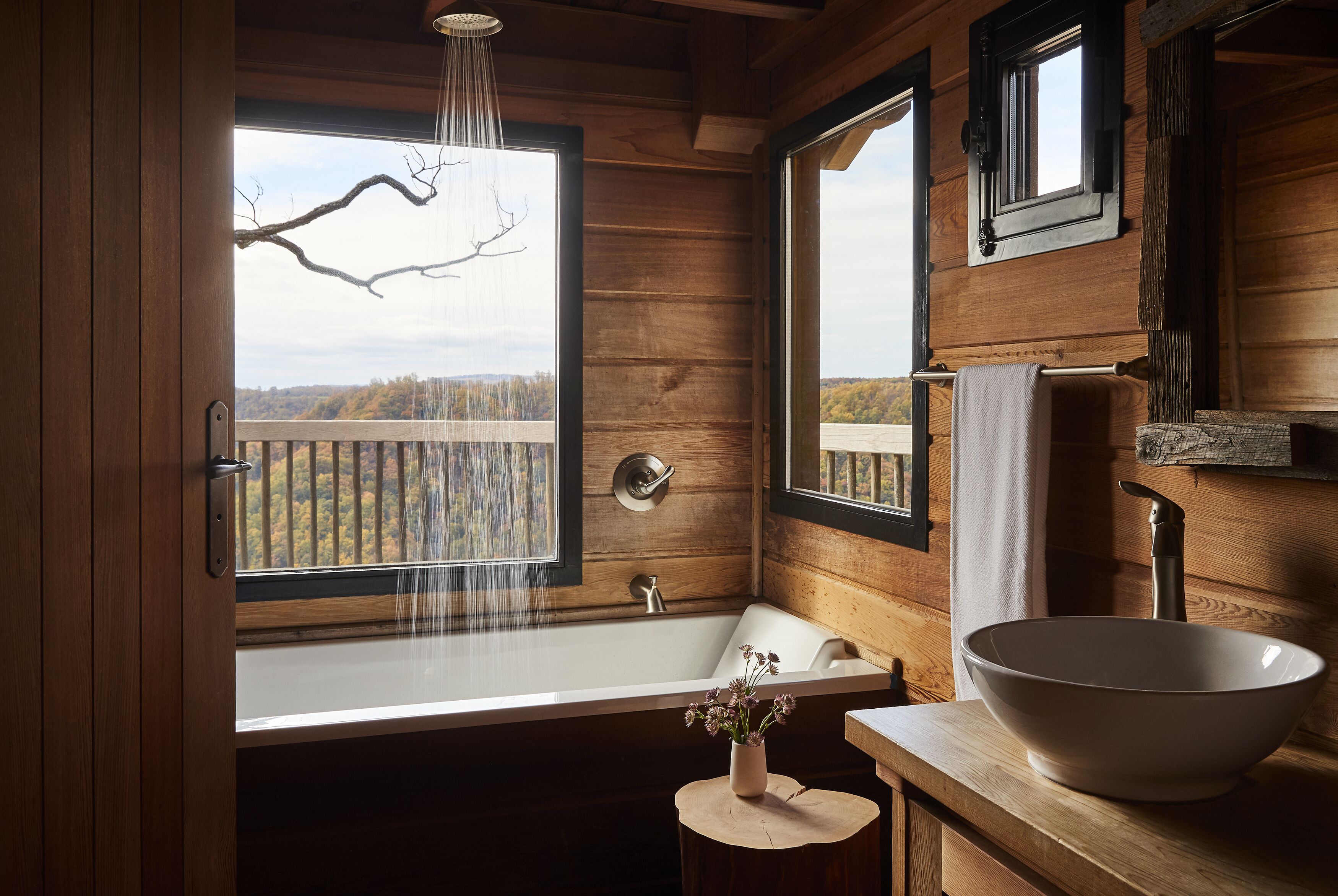 A wooden cabin bathroom with a large bathtub, a wooden vanity, and a view of a forest through a window.