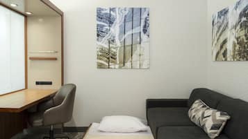 A modern hotel room with a desk, a gray office chair, a gray sofa, and two abstract paintings on the wall.