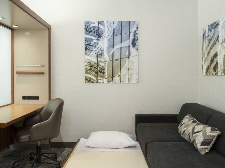 A modern hotel room with a desk, a gray office chair, a gray sofa, and two abstract paintings on the wall.