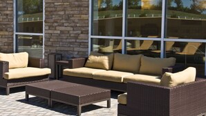 An outdoor seating area with wicker furniture and cushions, a stone wall, and large windows reflecting the surrounding landscape. - Courtyard by Marriott Owensboro (Owensboro)