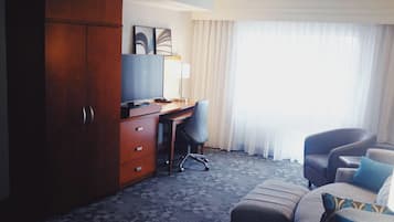 A hotel room with a wooden wardrobe, a desk with a chair, a sofa with cushions, and a large window with curtains.