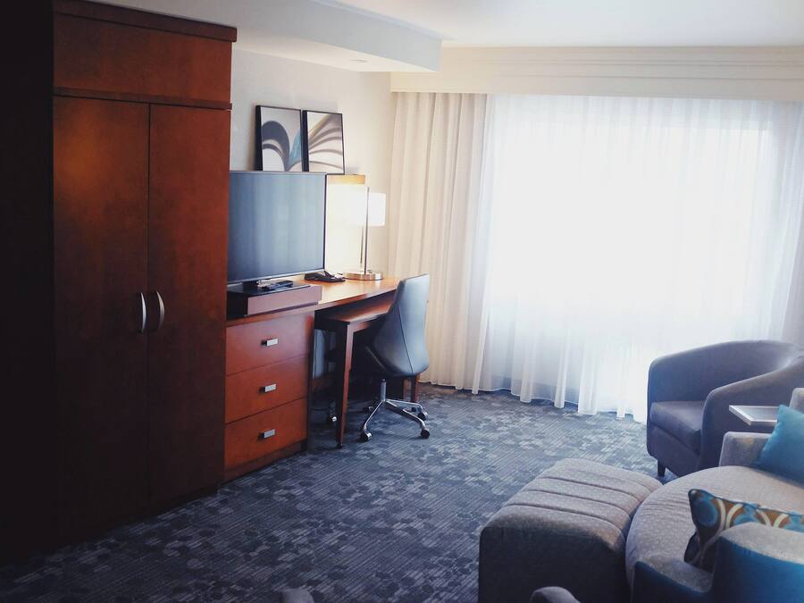A hotel room with a wooden wardrobe, a desk with a chair, a sofa with cushions, and a large window with curtains.