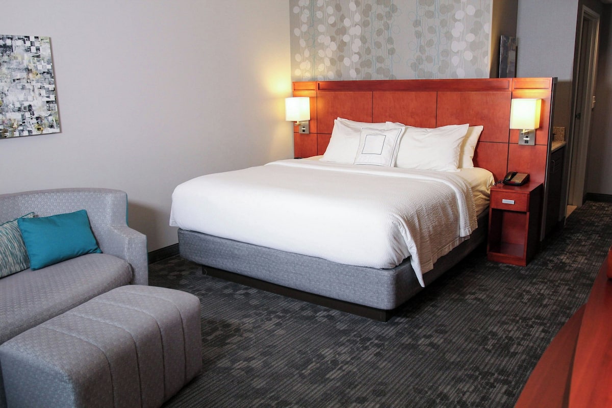Una habitación de hotel con una cama grande, dos lámparas de noche, una mesita de noche con un teléfono, un sofá gris con un cojín azul turquesa y una pared con diseño.