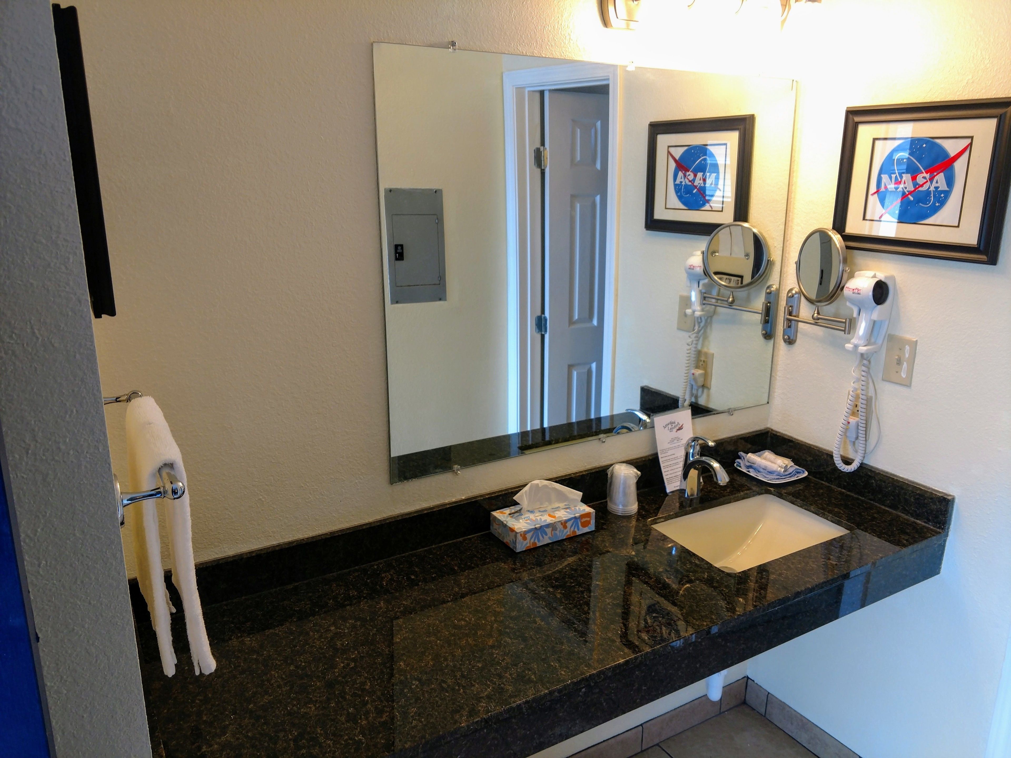 A hotel bathroom with a black granite countertop, a white sink, a hairdryer, and framed NASA and ACAM logos on the wall.