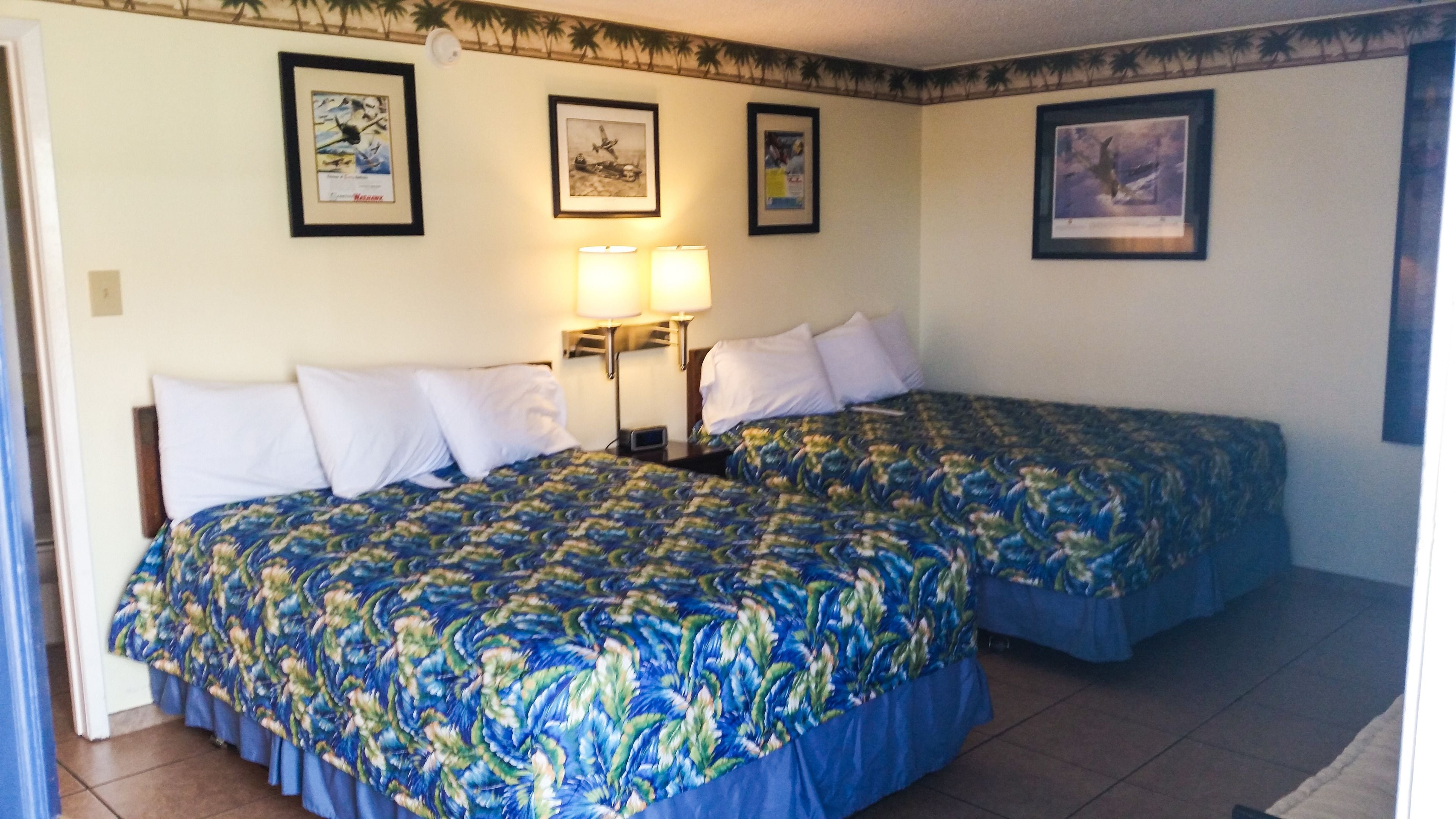 Une chambre d’hôtel avec deux lits, chacun recouvert d’une couvre-lit à motifs floraux, une table de chevet avec une lampe et des tableaux encadrés accrochés aux murs.