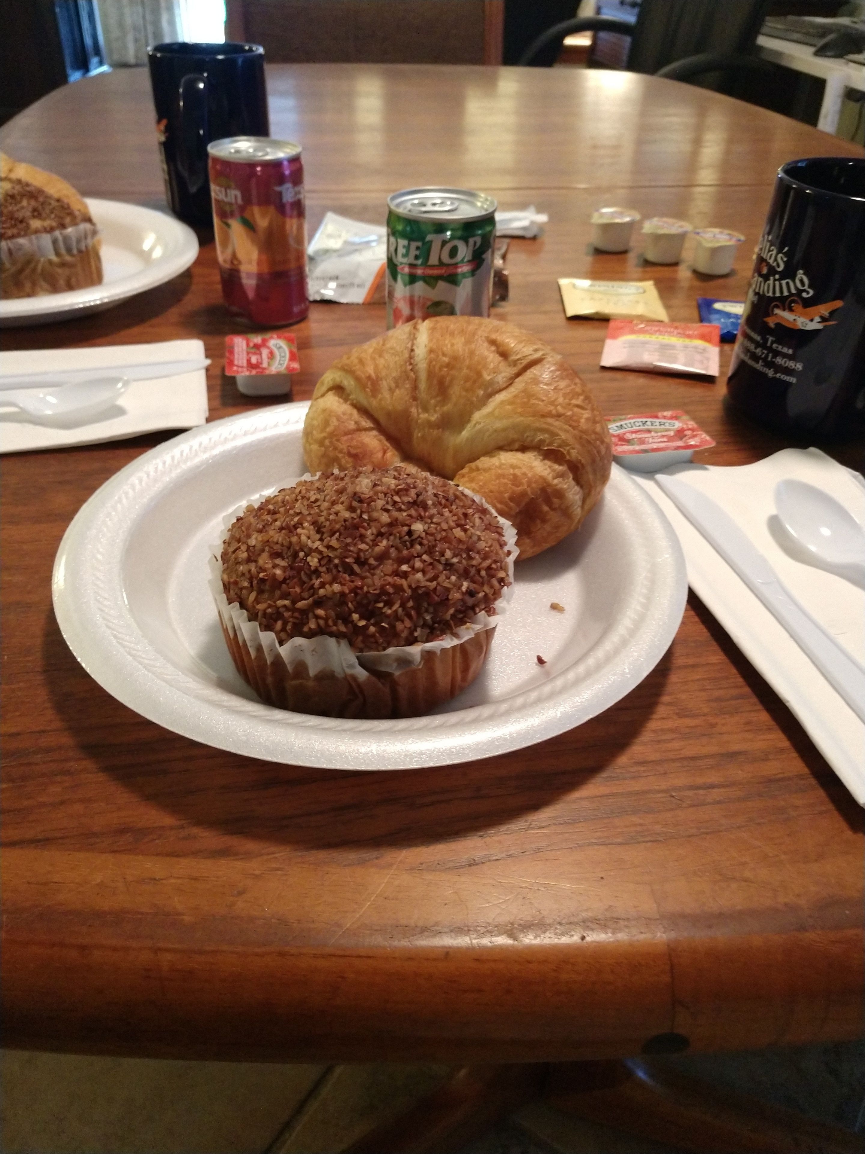 Une table avec un croissant et un muffin sur une assiette blanche, une canette de soda et une tasse noire.