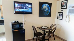Een kleine kamer met een aan de muur bevestigde televisie, een magnetron, een tafel met twee stoelen en afbeeldingen in lijsten aan de muur.