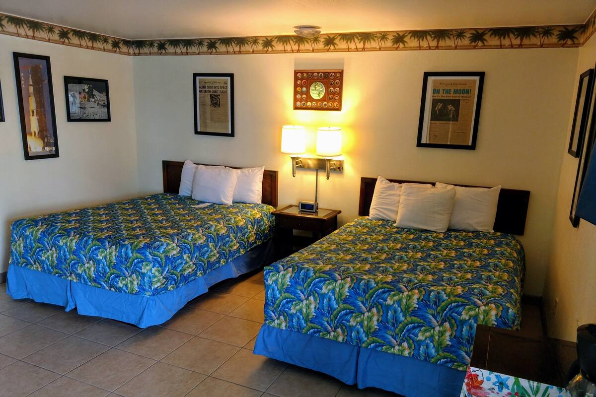 Une chambre d’hôtel avec deux lits, chacun recouvert d’une couvre-lit à motifs floraux, une table de chevet avec une lampe et des tableaux encadrés accrochés aux murs.