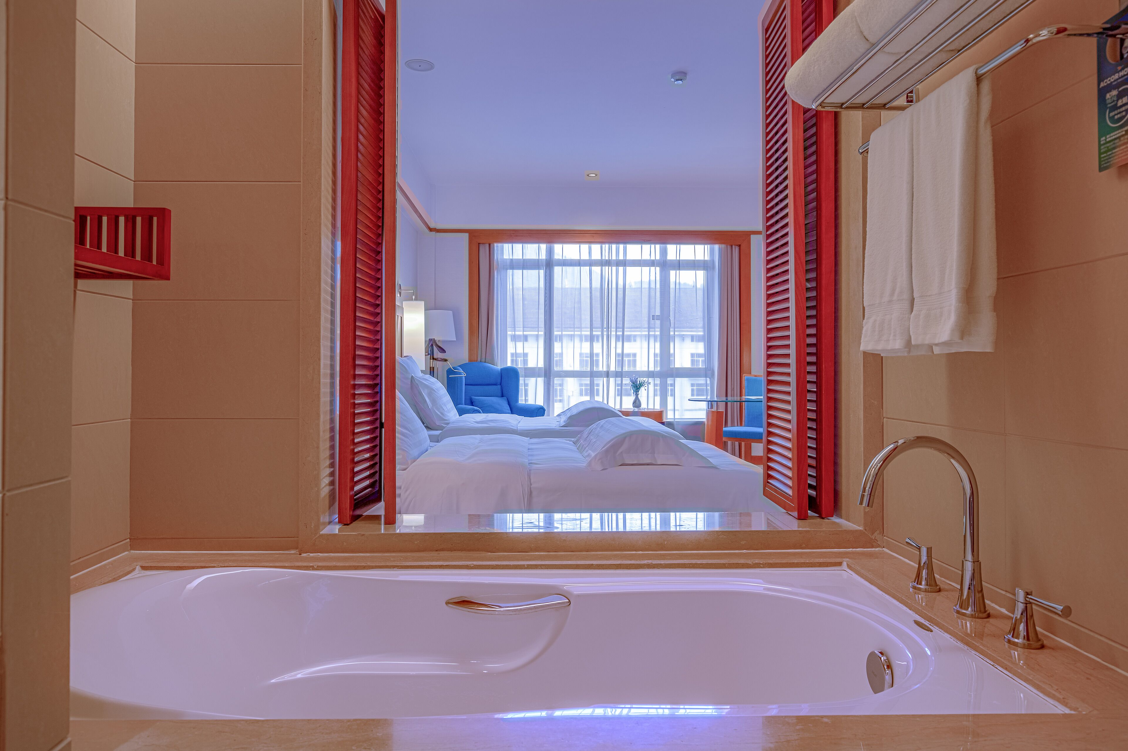 A modern bathroom with a large bathtub, a visible shower area, and a bed with blue bedding in the background.