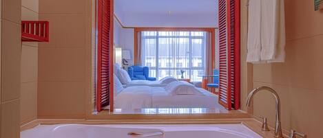A modern bathroom with a large bathtub, a visible shower area, and a bed with blue bedding in the background.
