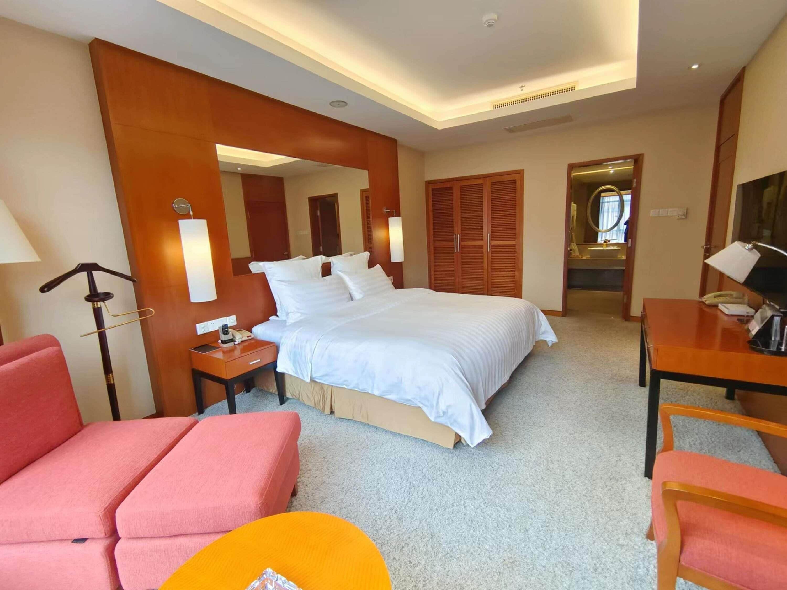 A hotel room with a bed, a desk, a chair, a lamp, a mirror, and a wardrobe.