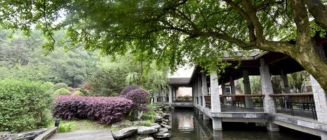 A wooden pathway leading to a stone bridge over a calm water body, surrounded by lush greenery and a traditional pavilion.