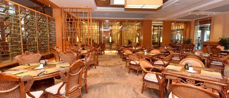 A hotel dining area with multiple tables and wicker chairs, a patterned carpet, and a wine rack in the background.