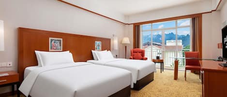A hotel room with two beds, a desk, a chair, and a large window with a mountain view.