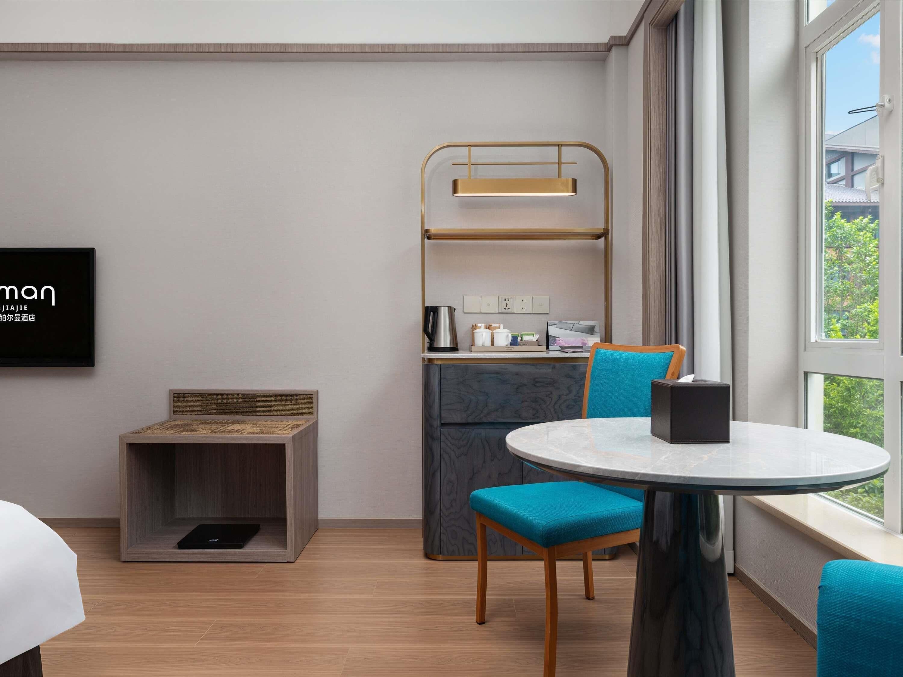 A modern hotel room with a round table, a sofa, a TV, a bookshelf, and a window with a view.