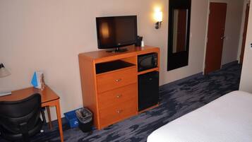 A hotel room with a bed, a desk with a computer, a television, a microwave, a trash bin, and a door.