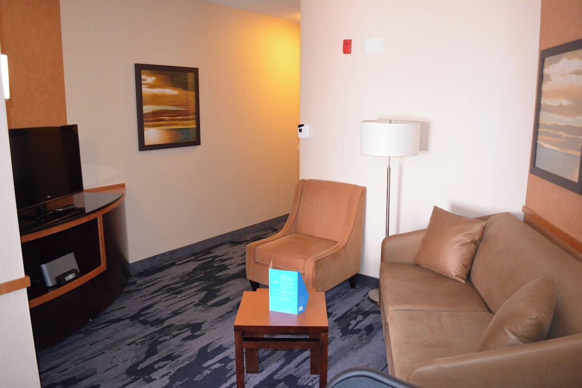 A hotel room with a sofa, a chair, a small table with a sign, a TV, and a lamp.