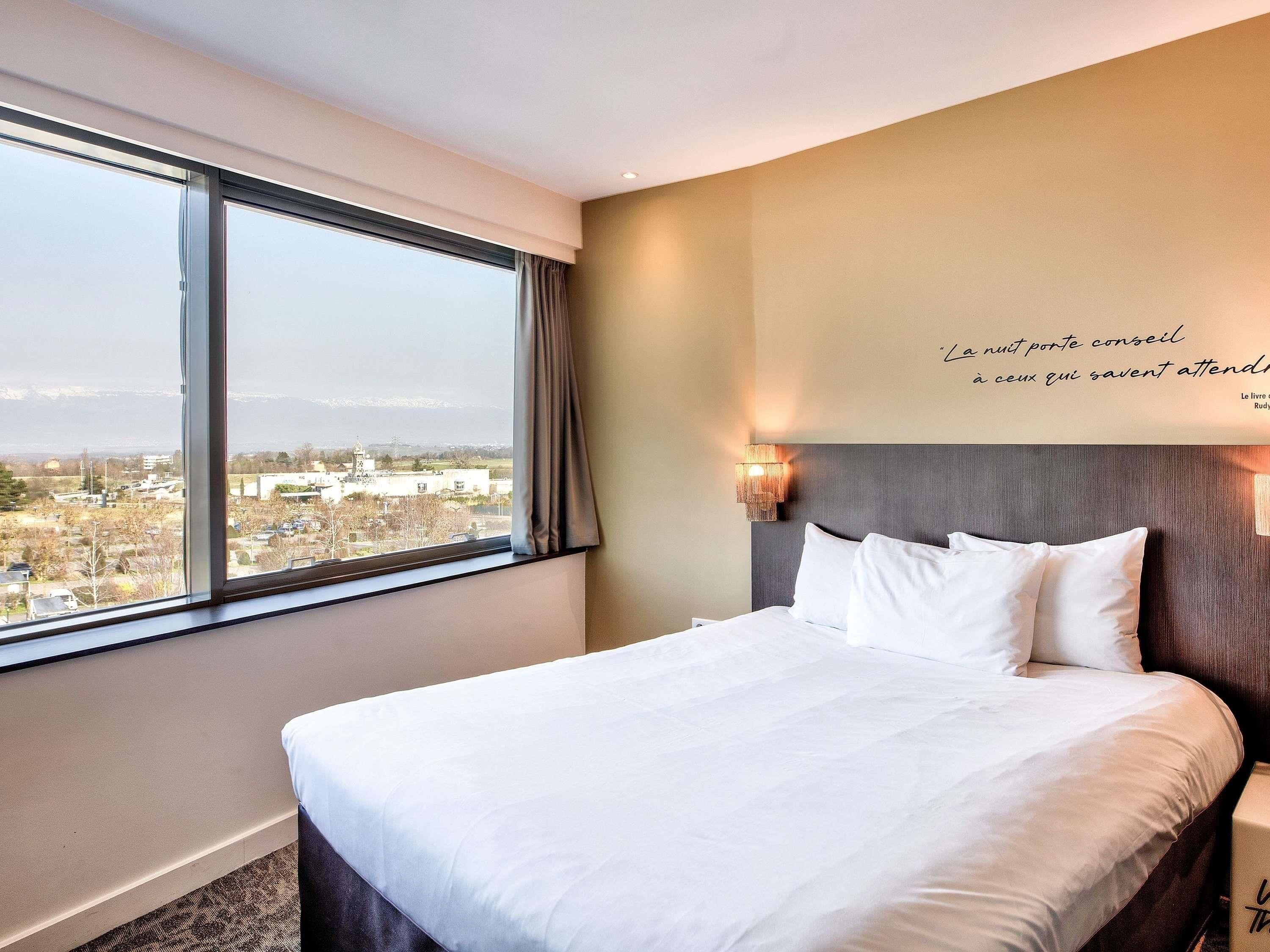 A hotel room with a large bed, a view of a cityscape through a window, and a wall-mounted light fixture.