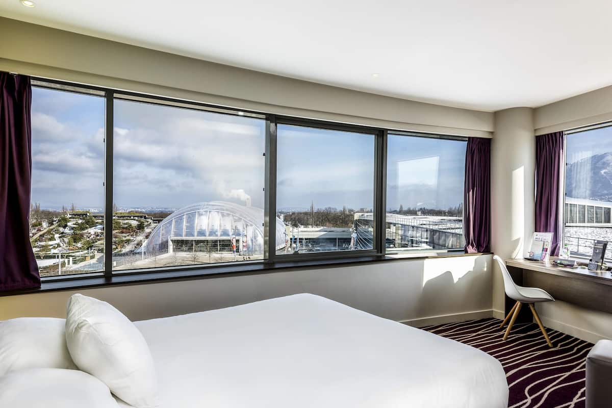 Une chambre d’hôtel avec un grand lit, un bureau, une chaise et une vue sur le paysage urbain, grâce à de grandes fenêtres.