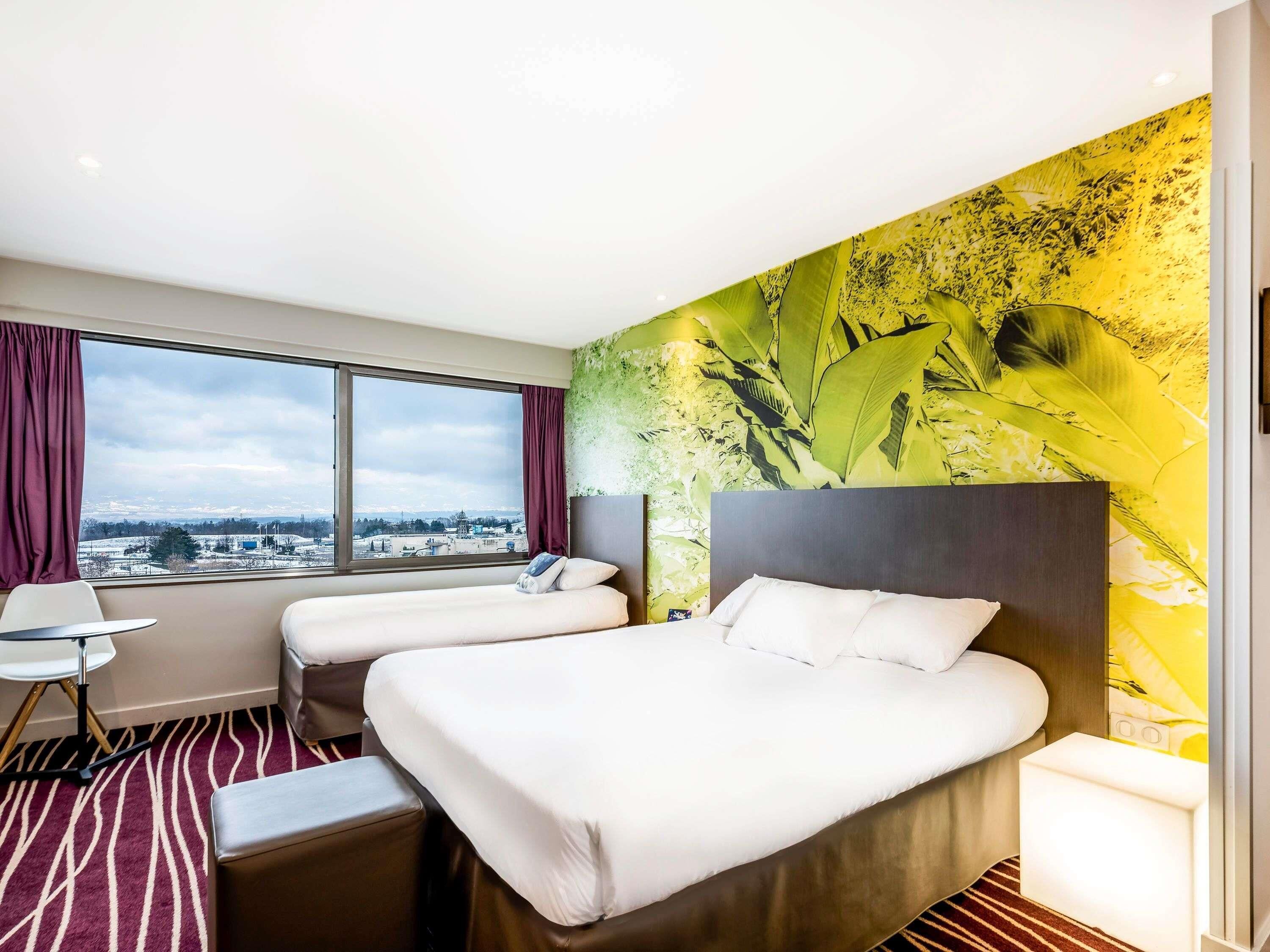 Habitación de hotel con dos camas, un escritorio, una silla y un ventanal con vista a la ciudad.