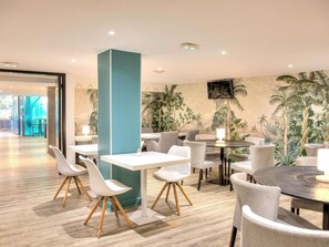 Un espace de restauration intérieur moderne, avec des tables et des chaises, une grande colonne de couleur turquoise et une fresque murale aux motifs tropicaux.