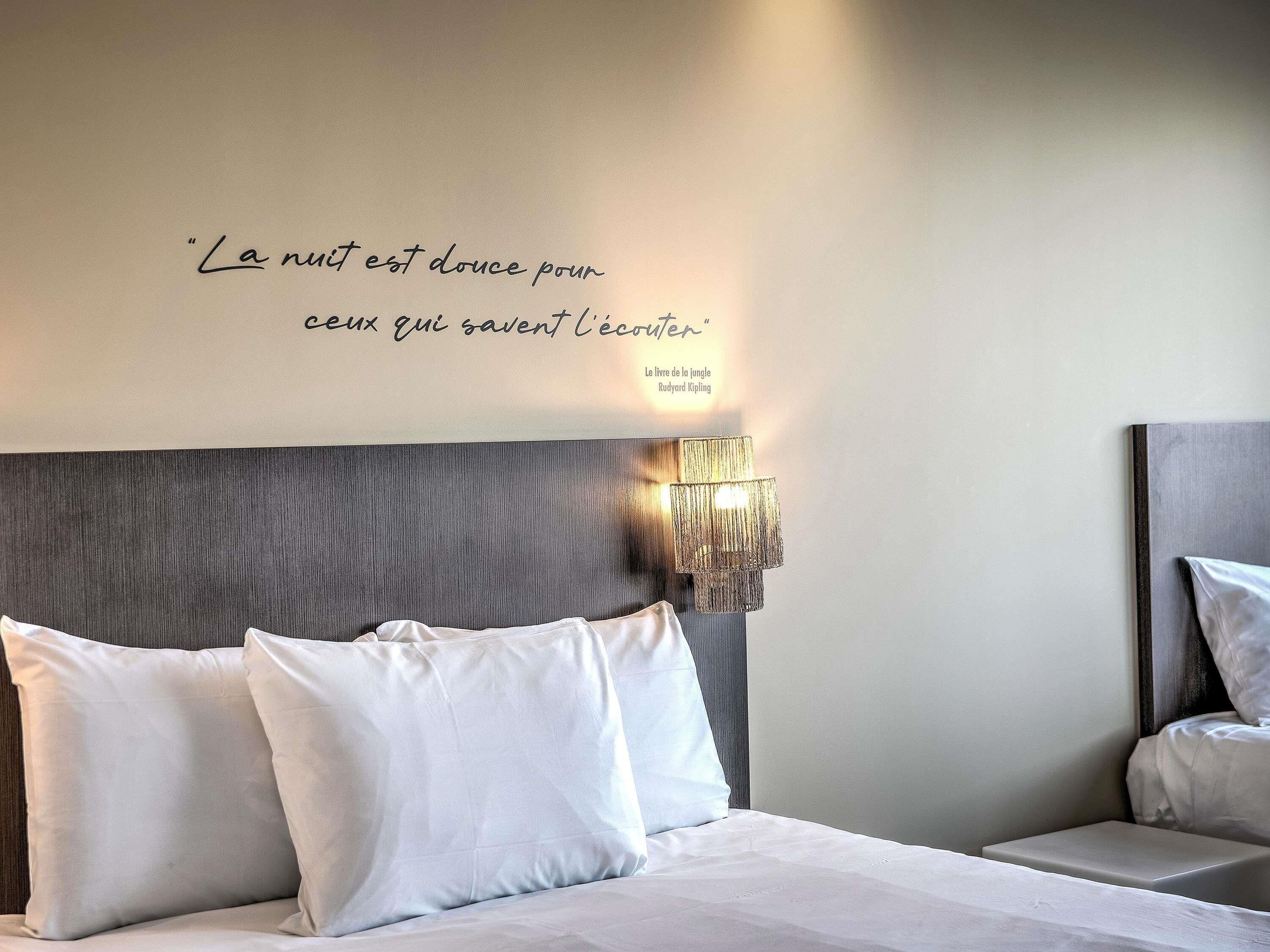 A neatly made bed with white linens and pillows, a wall-mounted lamp, and a quote in French.