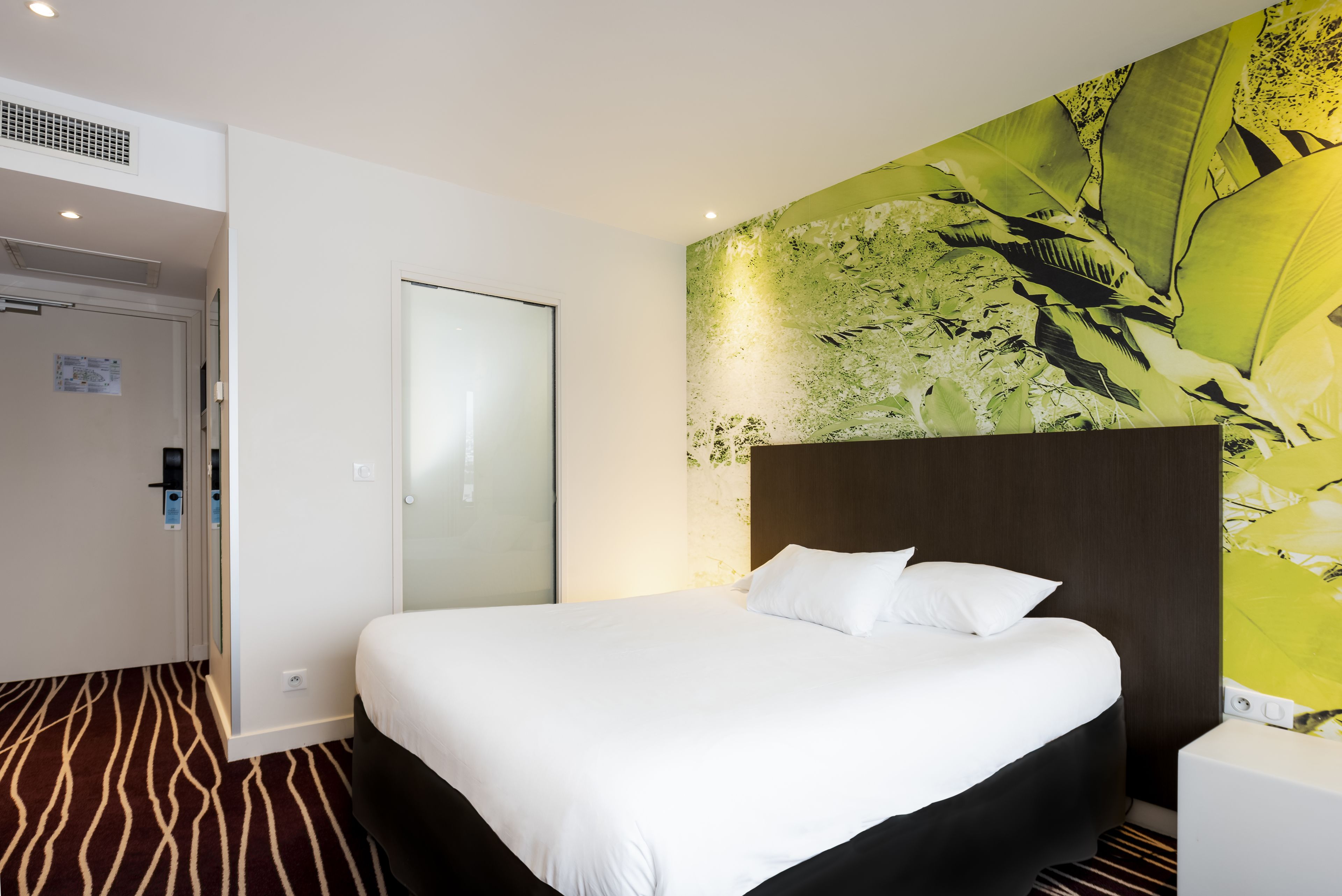 A hotel room with a large bed, a wooden headboard, a wall mural, a door with a glass panel, and a patterned carpet.