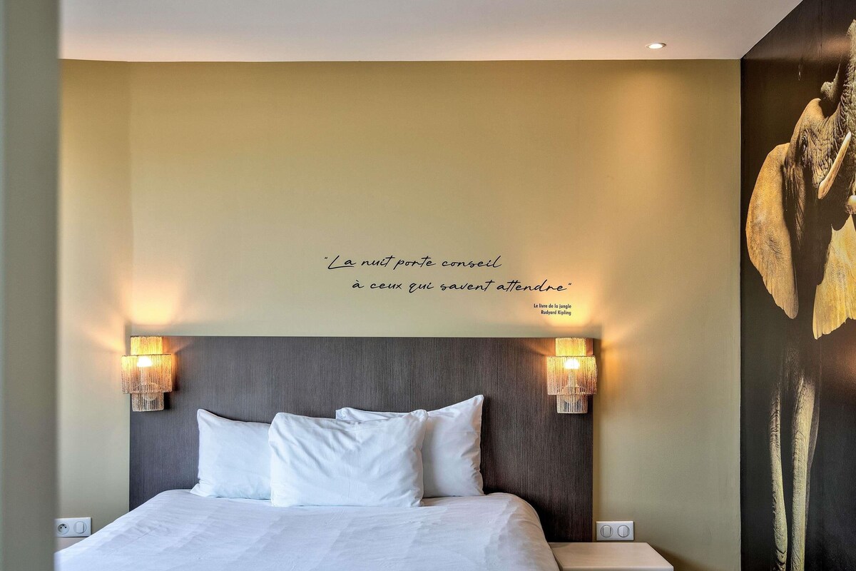Une chambre d’hôtel avec un lit, deux lampes fixées au mur, une citation décorative et un grand tableau représentant un éléphant.
