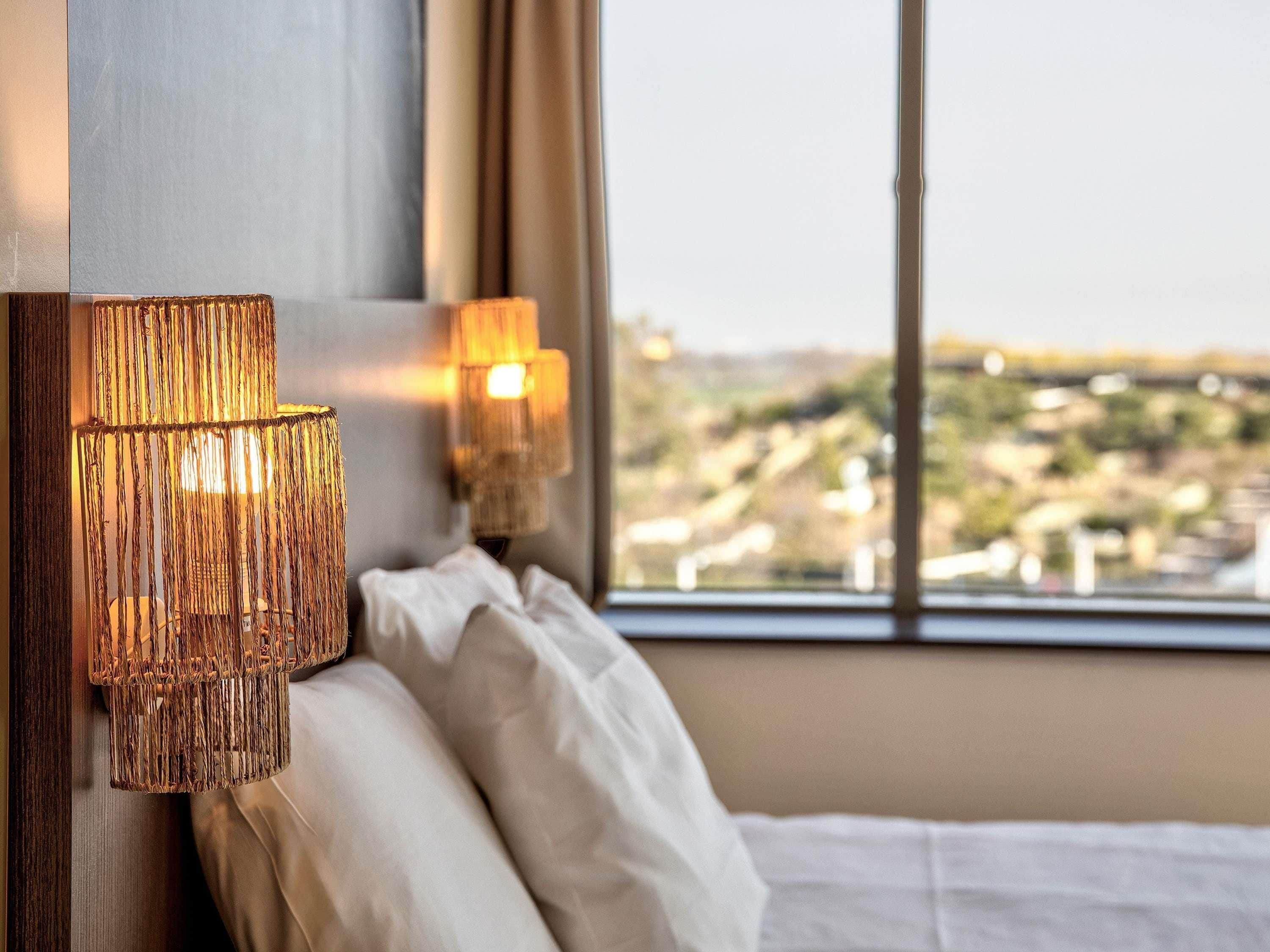 A hotel room with a bed, two wall-mounted lamps, and a view of a cityscape through a window.