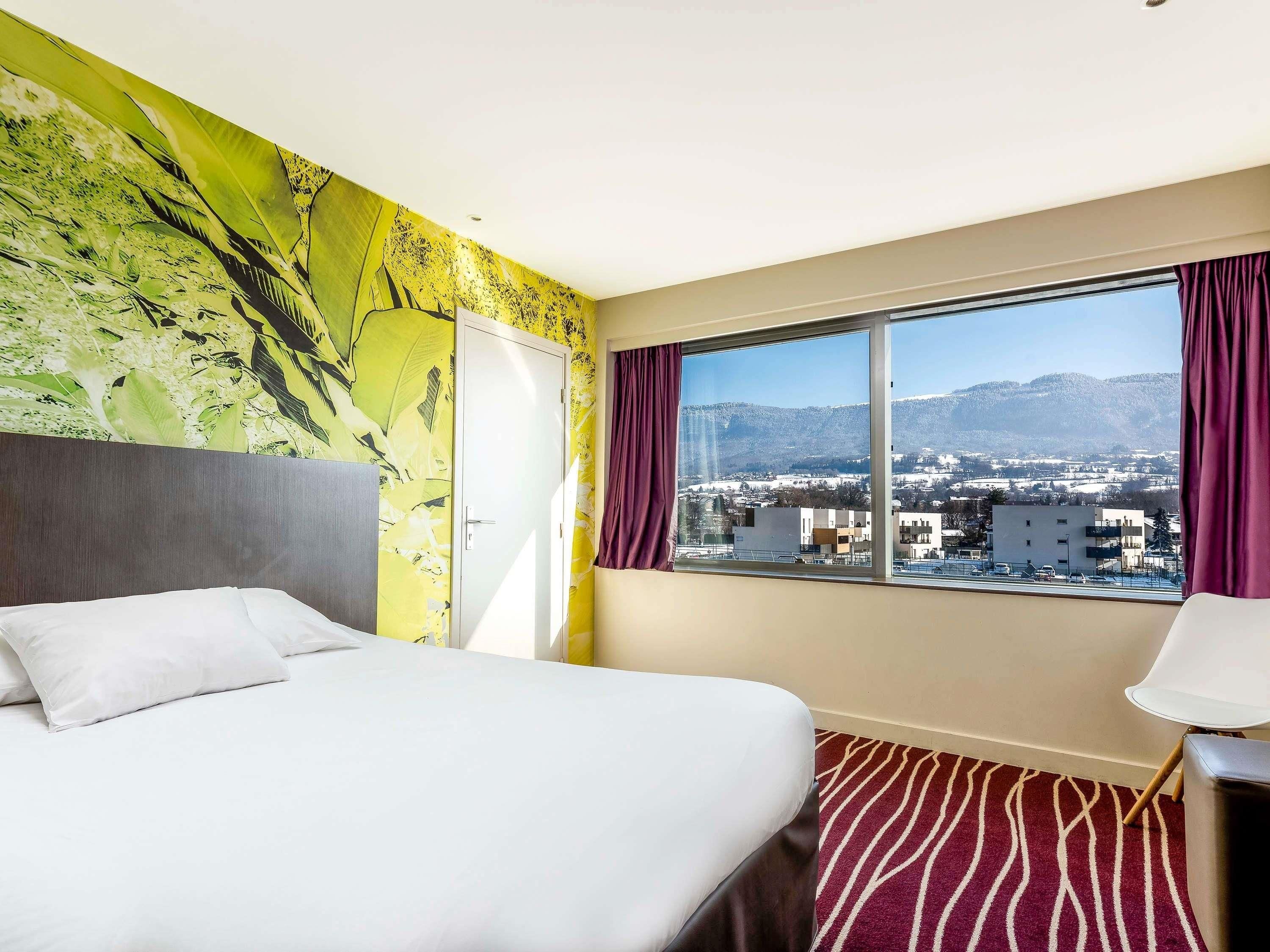 Une chambre d’hôtel avec un lit, une fenêtre donnant sur la ville et un mur orné d’une fresque aux motifs tropicaux.