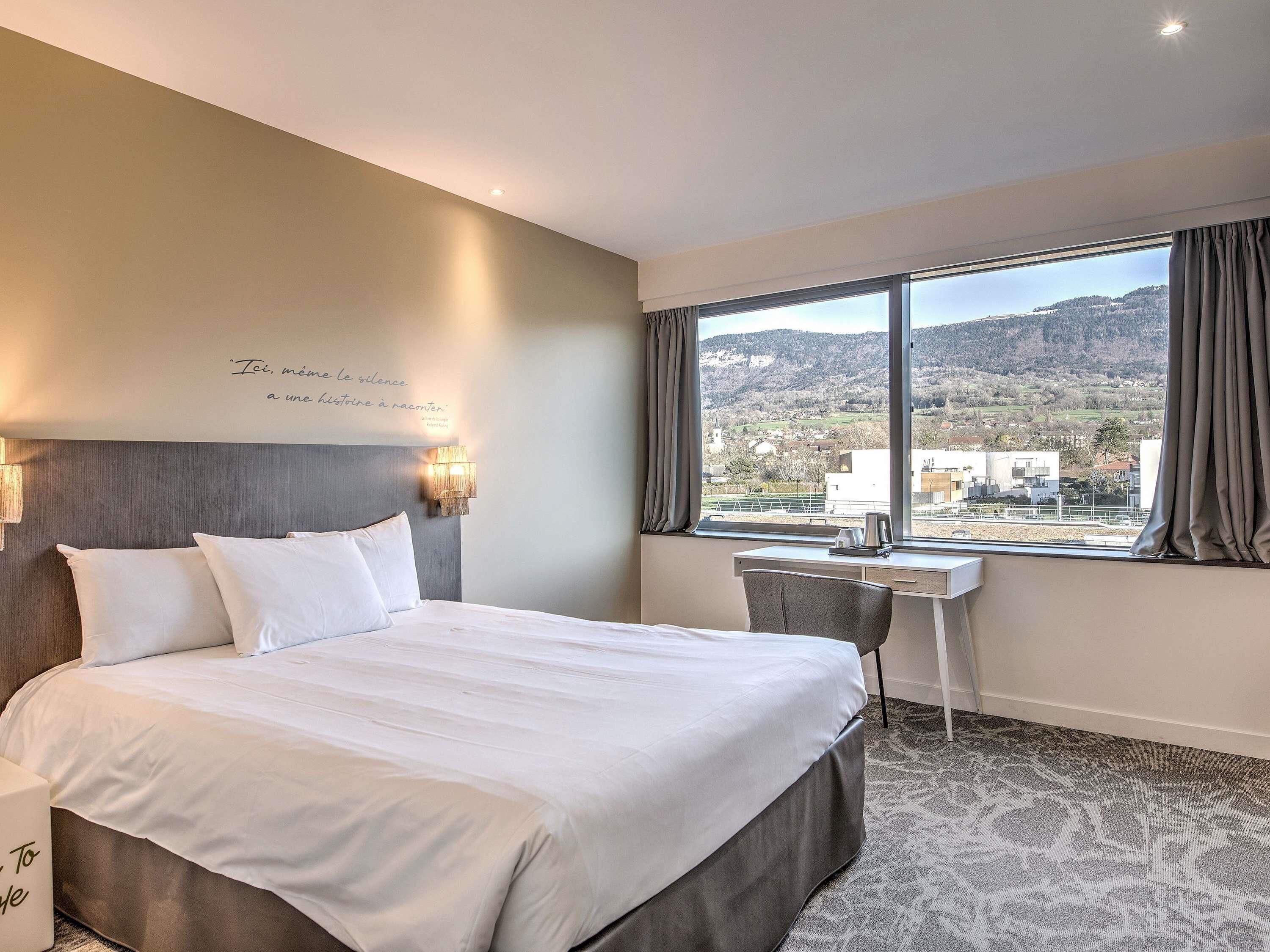 Habitación de hotel con cama, escritorio, silla, ventana que ofrece vistas a un pueblo y colinas, y una pared con una cita.