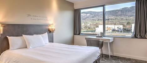 Habitación de hotel con cama, escritorio, silla, ventana que ofrece vistas a un pueblo y colinas, y una pared con una cita.