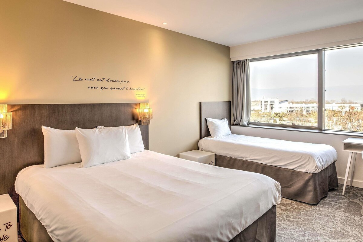 Une chambre d’hôtel avec deux lits, une grande fenêtre donnant sur la ville, une citation murale et un lustre.