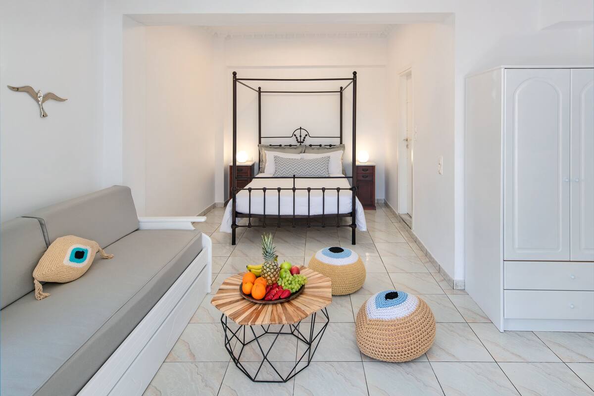A neatly arranged bedroom with a four-poster bed, a sofa with a crocheted pillow, a round table with fruit, and two woven poufs.