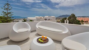 Una terraza en la azotea con mobiliario blanco moderno, una mesa pequeña con fruta y bebidas, y una vista panorámica del mar y las montañas.