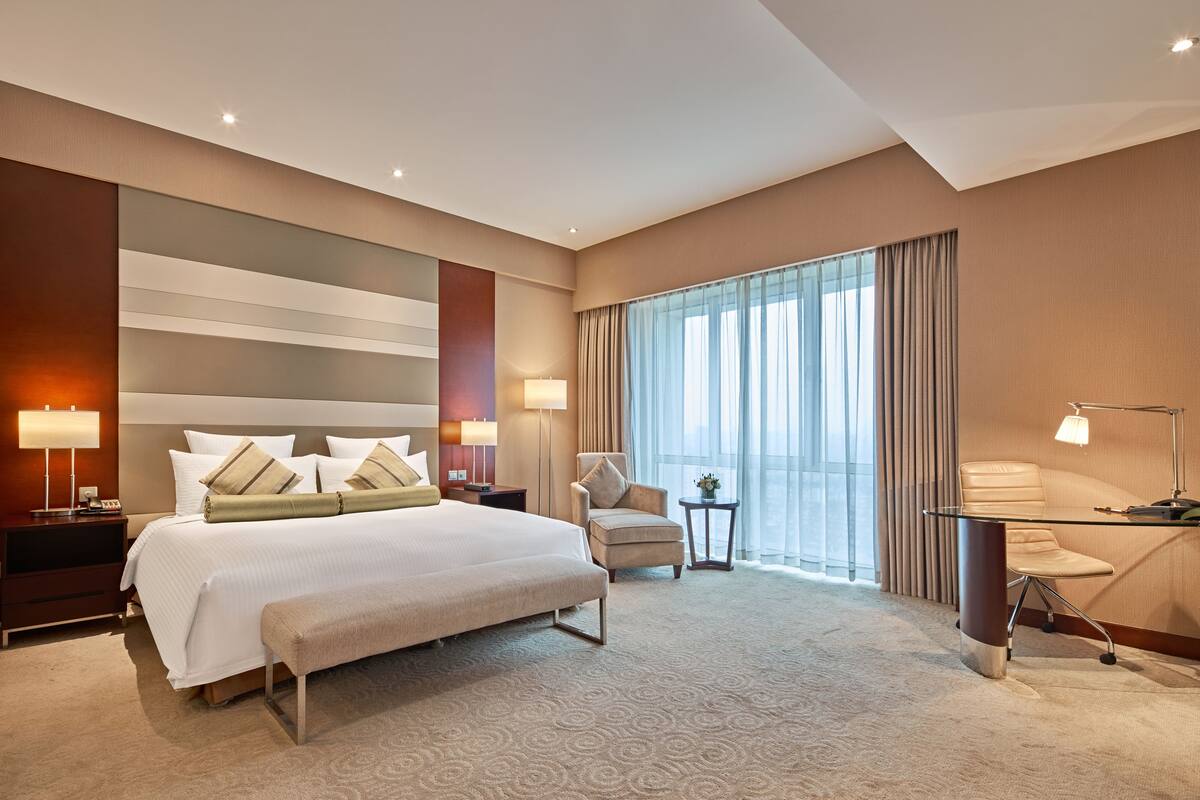 A modern hotel room with a large bed, a desk, a chair, and a view of the city through large windows.