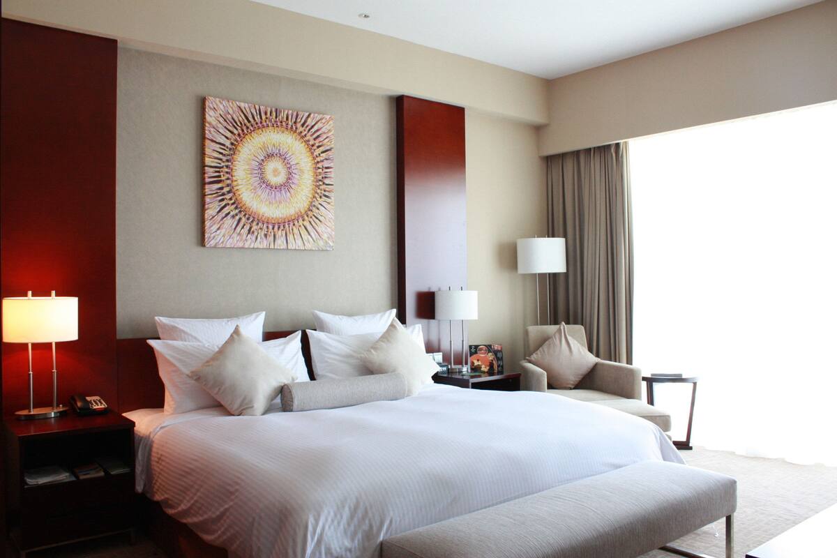 A hotel room with a large bed, a nightstand with a lamp, a wall-mounted artwork, and a view of the outdoors.