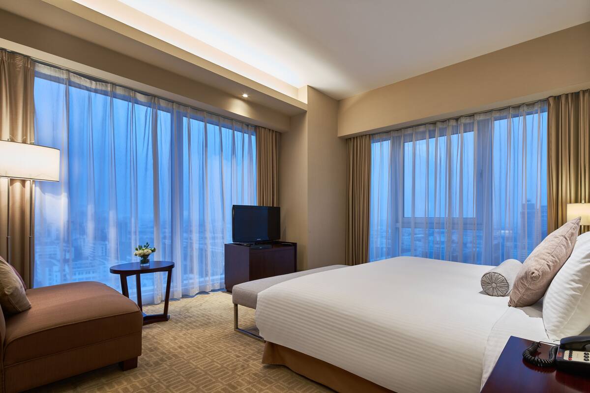 A hotel room with a large bed, a TV on a stand, a small table with a vase, and a view of the city through sheer curtains.