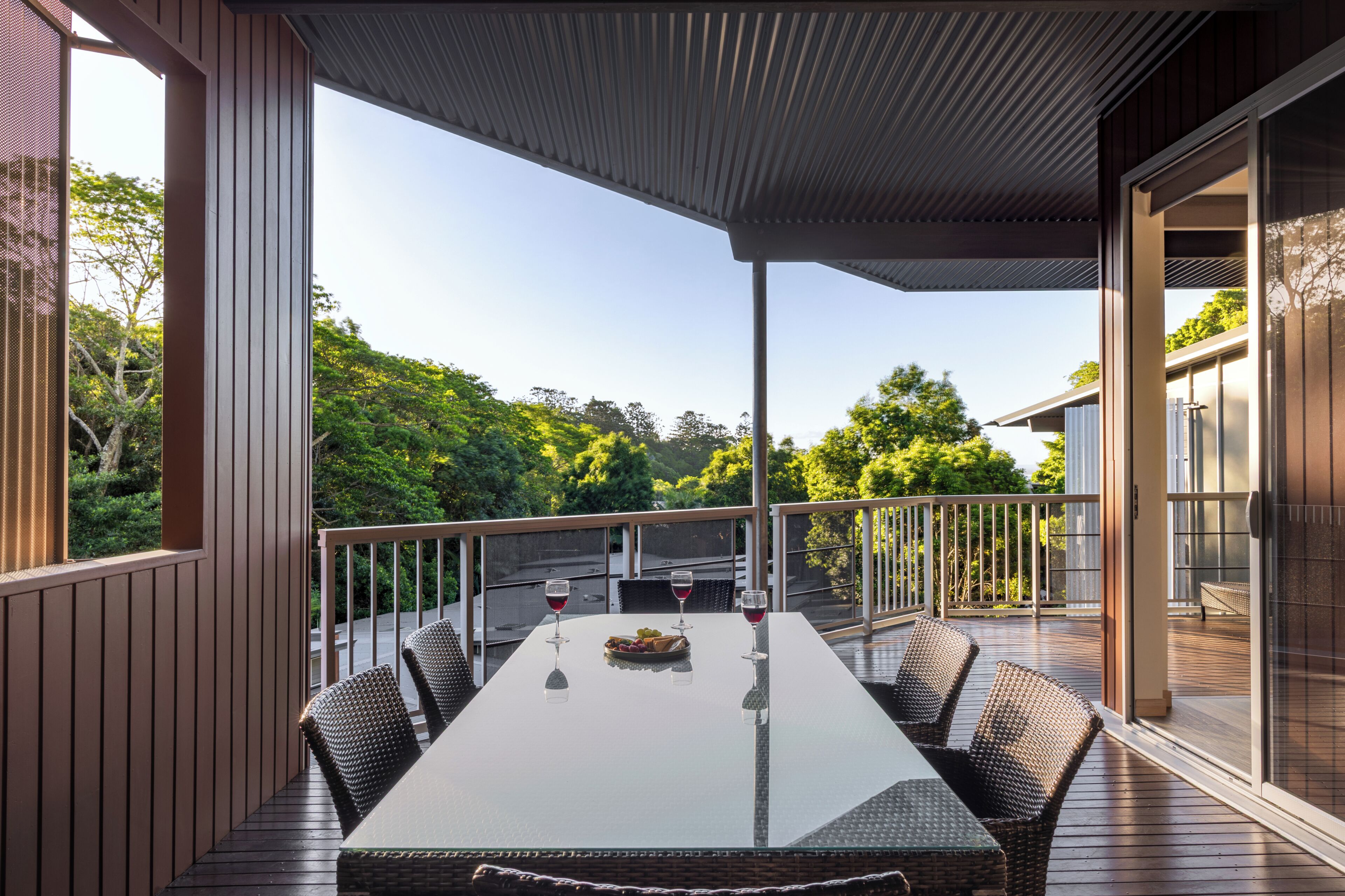 A covered outdoor dining area with a table, chairs, and wine glasses, overlooking a lush green landscape.