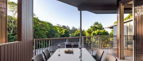 A covered outdoor dining area with a table, chairs, and wine glasses, overlooking a lush green landscape.