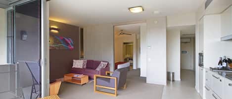 A modern living room with a sofa, chairs, a coffee table, and a small wooden table. There is a kitchen area with cabinets and a countertop visible.