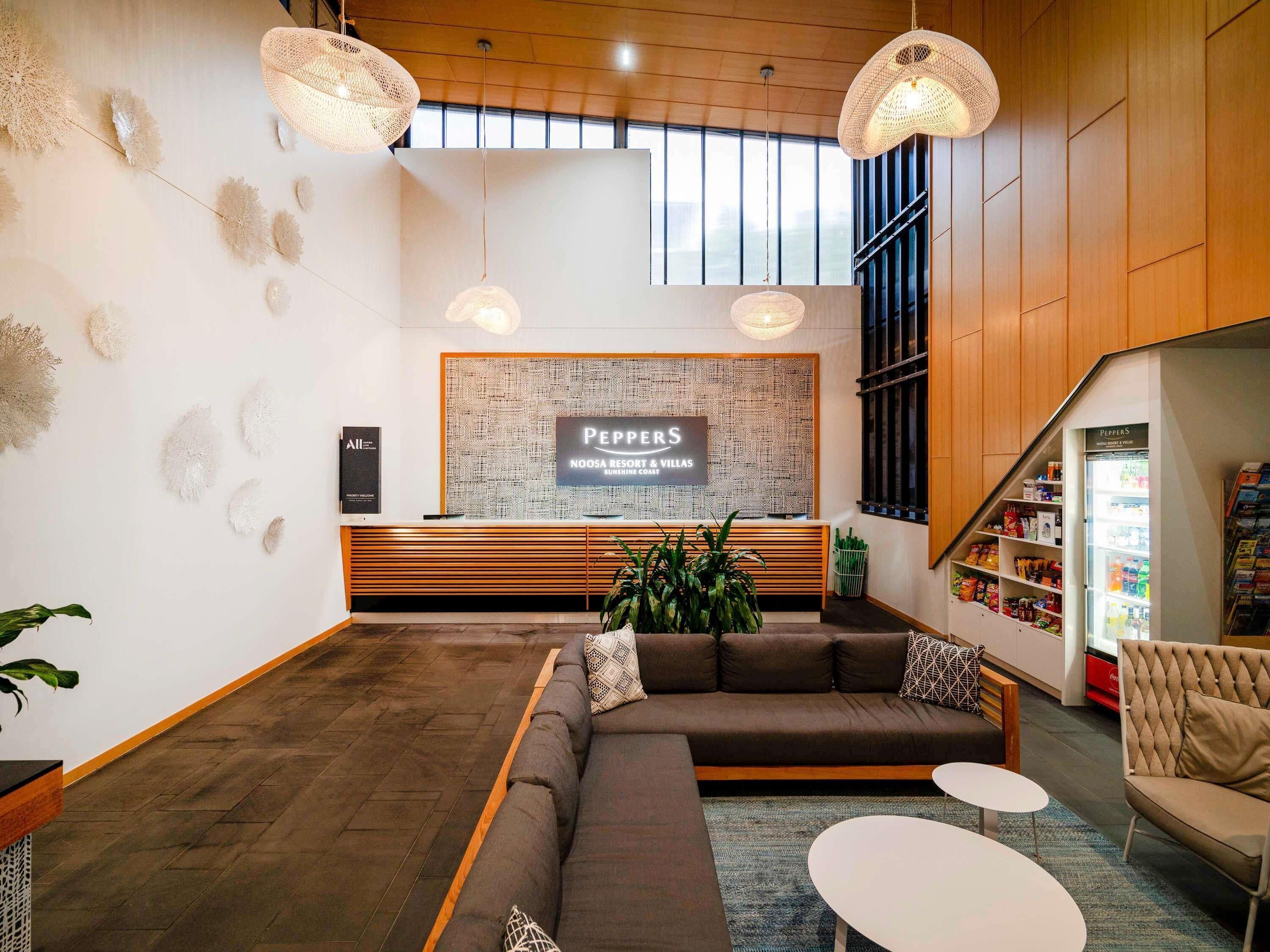 A reception area with a sign for 'Peppers Noosa Resort & Villas,' a reception desk, a sofa, two small tables, and a refrigerator stocked with beverages.