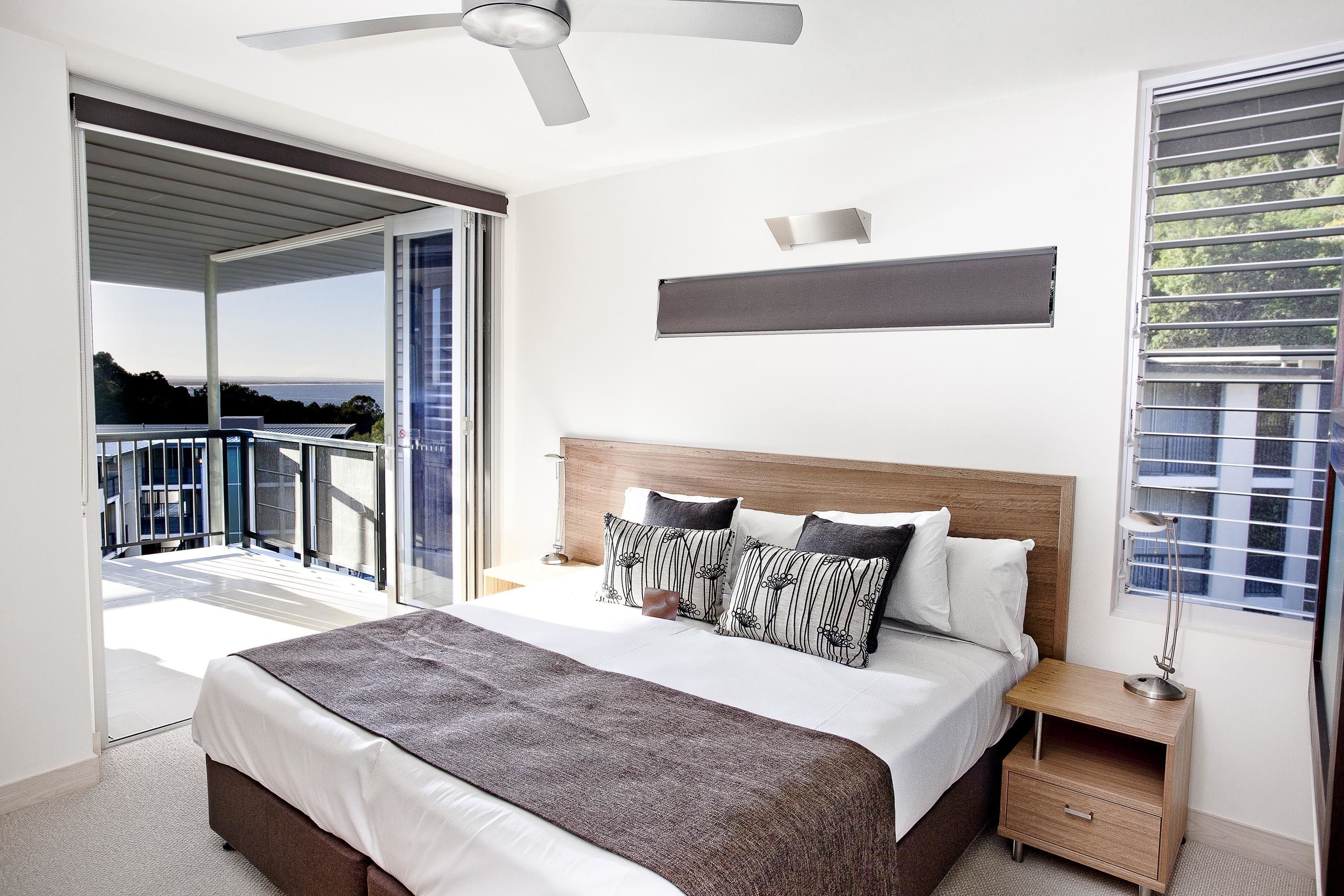 Une chambre avec un grand lit, une table de chevet, un ventilateur de plafond et une vue sur l’extérieur.